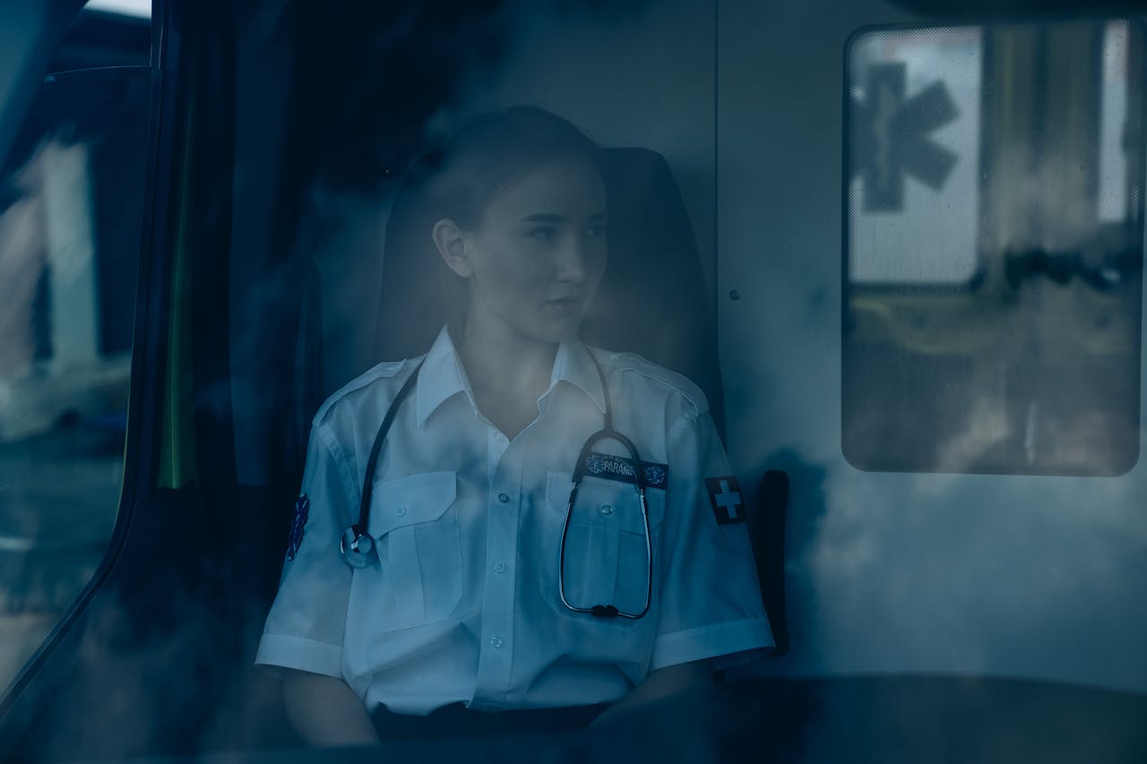 A thoughtful paramedic sits inside an ambulance, wearing a uniform and stethoscope.
